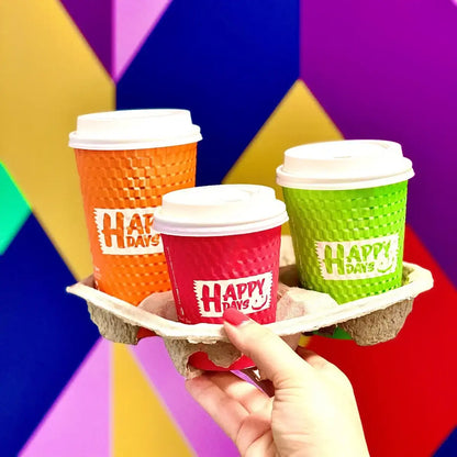 Three colorful coffee cups with 'Happy Days' branding on a tray against a multicolored geometric background.