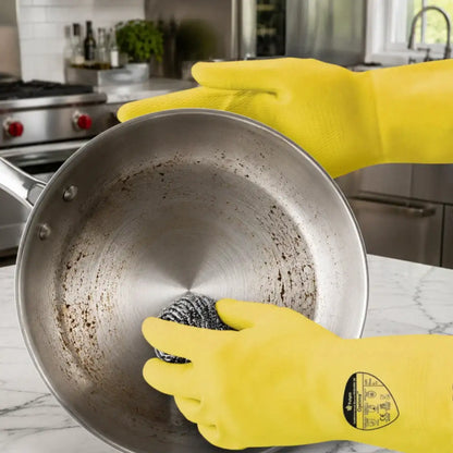 Durable Household Yellow Rubber Gloves being worn by a person inside a dirty stainless steel pan, scrubbing with a steel wire sponge on a marble countertop in a kitchen.