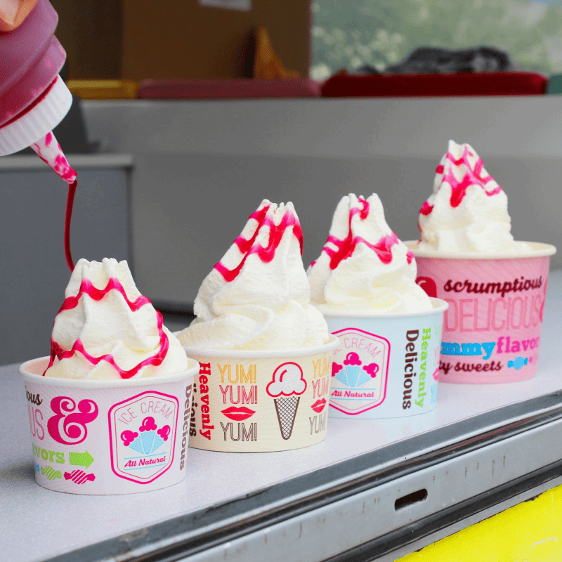Ice cream cups with colorful labels being filled with white ice cream and strawberry sauce.