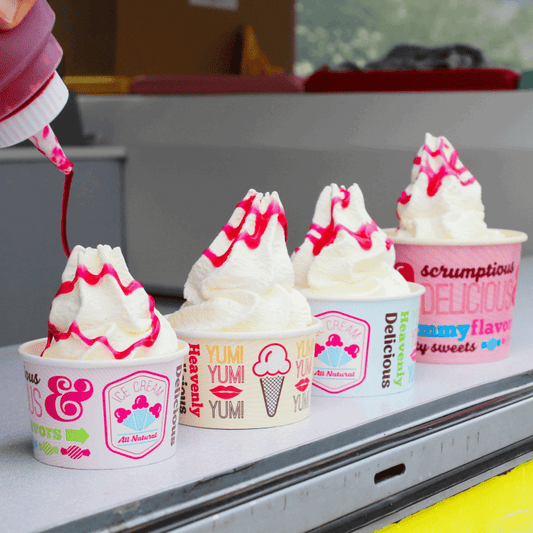 Ice cream cups with colorful labels being filled with white ice cream and strawberry sauce.